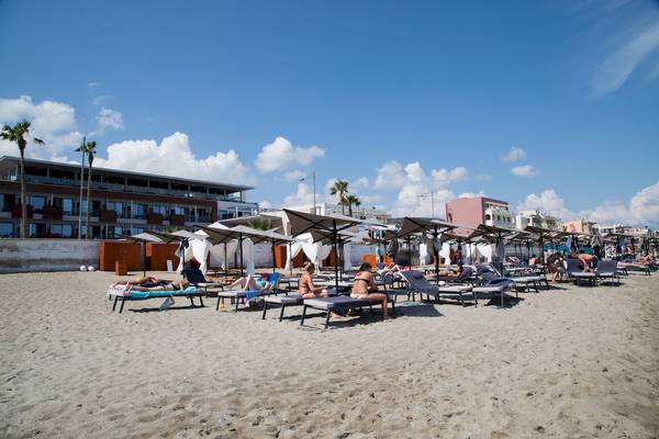 Strand mit Sonnenliegen und Gästen im Schatten von Sonnenschirmen am Ikones Seafront Luxury Suites Hotel.