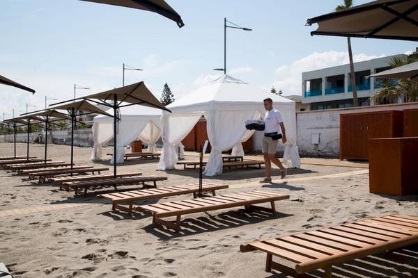 Mann auf dem Strand der Ikones Seafront Luxury Suites, Geht mit Gepäck durch Loungestühle und Pavillons.