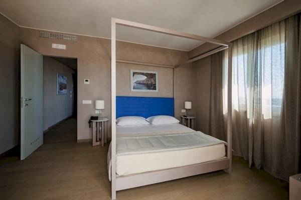 Schlafzimmer im Cala Ponte Hotel mit einem Bett mit blauem Kopfteil, zwei Nachttischen und Lampen.