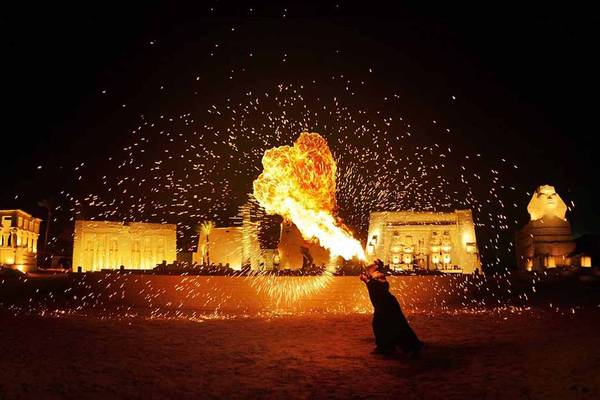 Eine Person spuckt Feuer in der Nacht vor alten Gebäuden und einer Sphinx-Statue.
