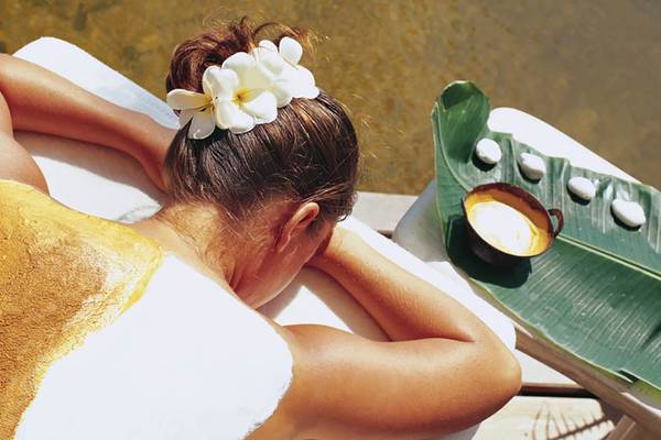 Rückenansicht einer Frau im Porto Elounda Golf & Spa Resort, die eine goldene Körperbehandlung genießt, mit einer weißen Blume im Haar und einem grünen Blatt neben einer Tasse.