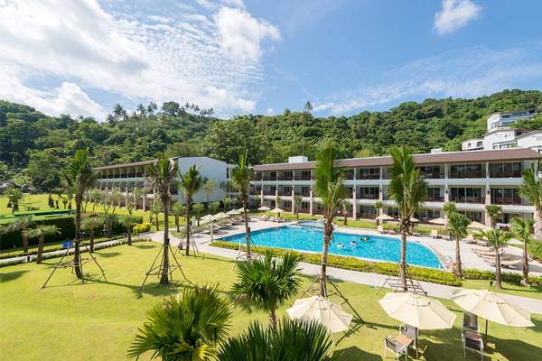 Außenansicht des Katathani Phuket Beach Resorts mit Swimmingpool, Palmen und Hügel im Hintergrund.