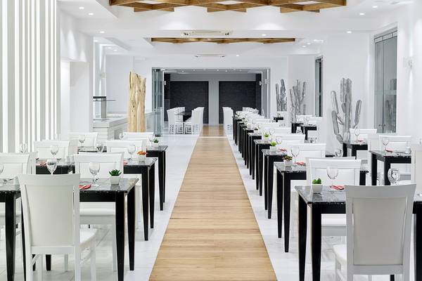 Innenansicht des Restaurants im Dimitra Beach Hotel & Suites mit weißen Tischen und Stühlen, einem hölzernen Gang und Kaktus-Dekorationen.