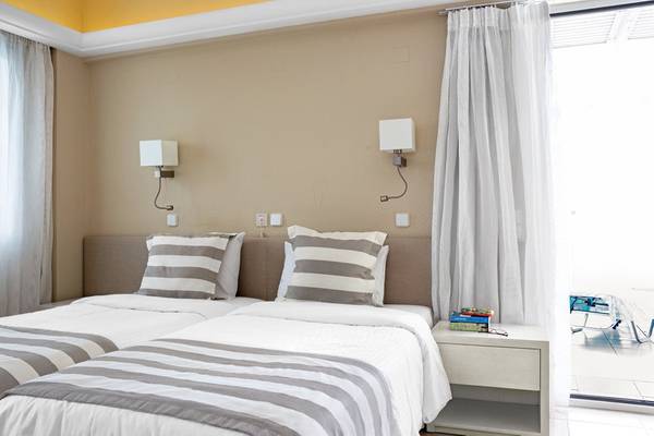 Zwei Betten im Hotelzimmer von Pearle Beach Resort & Spa, beide mit grau-weiß gestreiften Kissen und Decken, neben einem Nachttisch mit Büchern und einer Lesebrille, und einer offenen Tür mit Blick auf einen Balkon.