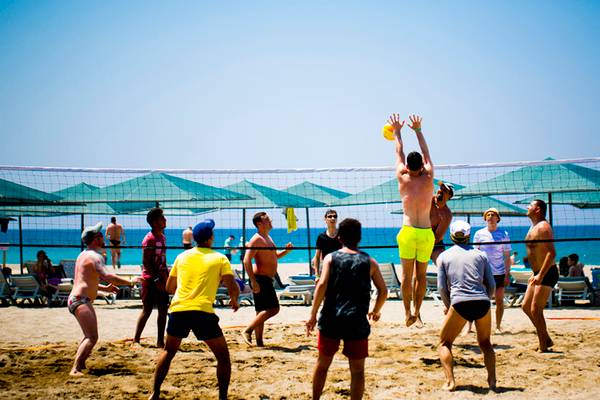 Mehrere Männer spielen Volleyball auf einem Strand mit einem Netz und Sonnenschirmen im Hintergrund.