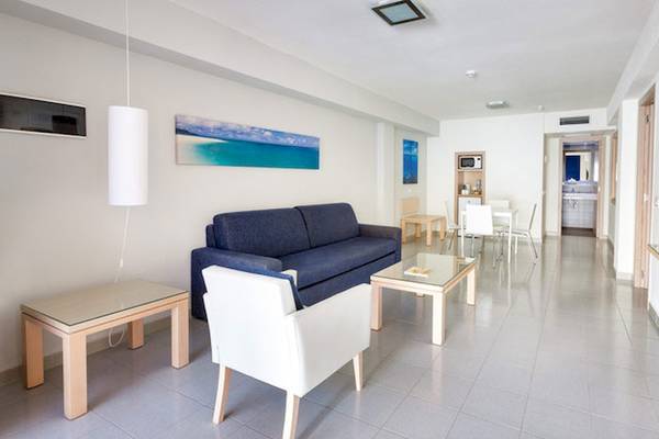 Inneneinrichtung des Tagoro Family & Fun Costa Adeje mit einem blauen Sofa, weißem Stuhl, Glastisch und Fernseher an der Wand.