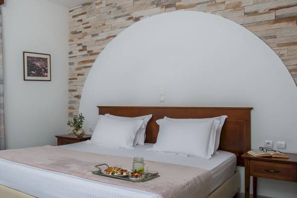 Schlafzimmer mit Frühstücksplatte im Naxos Palace Hotel, Bett mit Kissen, Frühstücksplatte mit Essen, Nachttisch mit Buch und Brille, Steinwand im Hintergrund.