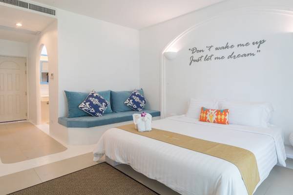 Schlafzimmer im Ocean Breeze Resort Khao Lak mit weißem Bett, blauem Sitzkissen, Wandspruch und Nachttischlampe.
