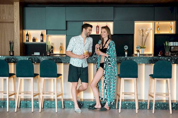 Ein Mann und eine Frau stehen lächelnd in einem Barbereich des Mitsis Rodos Village Beach Hotel & Spa und halten Cocktails in den Händen.