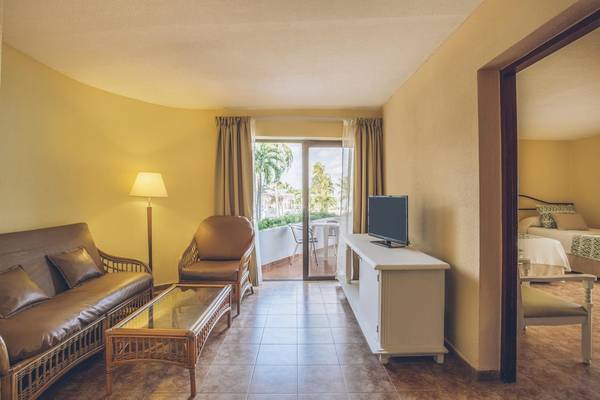 Hotelzimmer im Iberostar Origin Bella Costa mit gelben Wänden, einer Couch, einem Tisch und einem Fernseher.