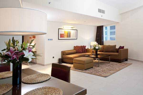 Das Wohnzimmer des Grand Millennium Al Wahda Hotels mit einem großen Sofa, einem Tisch, Blumen und einer Lampe.