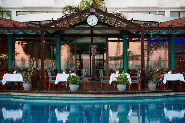 Pool-Lounge-Bereich im Hotel Louis Ledra Beach mit Tischen und Stühlen, Pflanzen und einer Uhr.