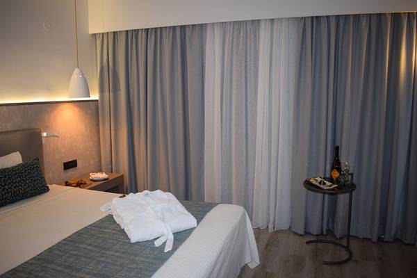 Hotelzimmer im Minos Mare mit Bett, grauen Vorhängen und Nachttisch mit Flasche und Gläsern.