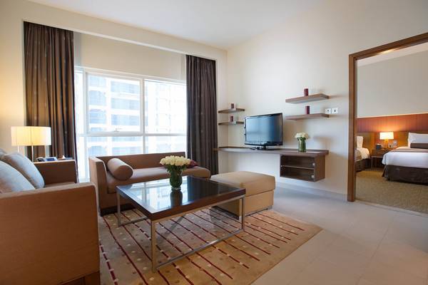 Innenansicht der Suite im Grand Millennium Al Wahda Hotel mit Wohnzimmer, Fernseher und Schlafbereich.