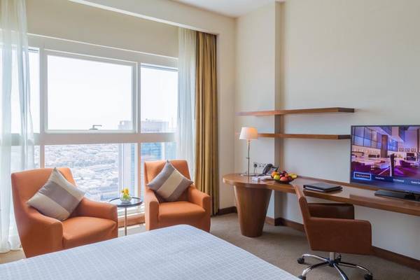 Innenansicht eines Hotelzimmers im Grand Millennium Al Wahda mit Schreibtisch, Fernseher und zwei Sesseln.