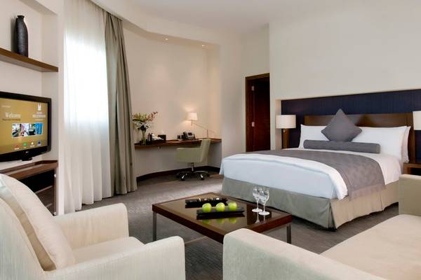 Innenansicht einer Suite im Grand Millennium Al Wahda Hotel mit Bett, Schreibtisch und Fernseher.