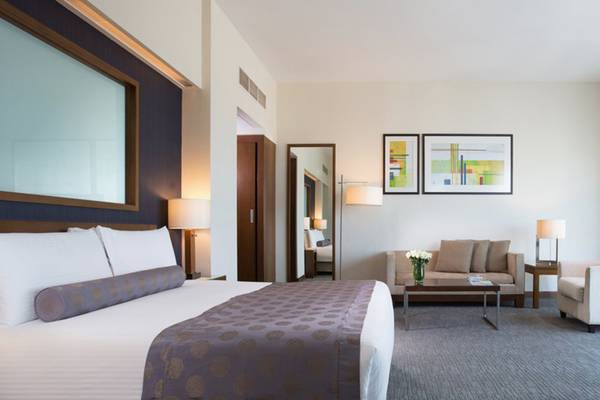 Innenansicht einer Suite im Grand Millennium Al Wahda Hotel mit Bett, Sofa, Tisch und Fenstern.
