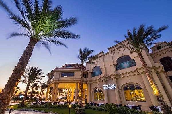 Außenansicht des Rixos Sharm El Sheikh Hotels bei Sonnenuntergang mit Palmen und beleuchtetem Eingang.