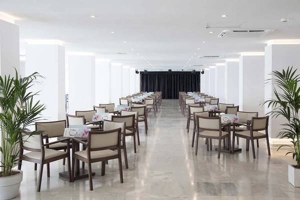 Innenansicht des Speisesaals im Alua Leo Hotel mit geräumigen weißen Wänden, braunen Stühlen und Tischen, sowie grünen Pflanzen in Töpfen.