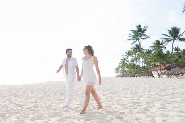 Ein Paar, das Hand in Hand auf einem weißen Sandstrand läuft, mit Palmen und Gebäuden im Hintergrund.
