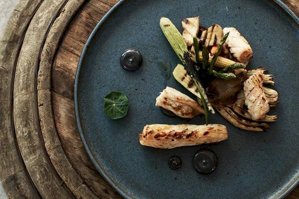 Ein Gericht auf einem blauen Teller mit Holzuntergrund, bestehend aus gegrilltem Fisch, Gemüse und schwarzen Soßen.