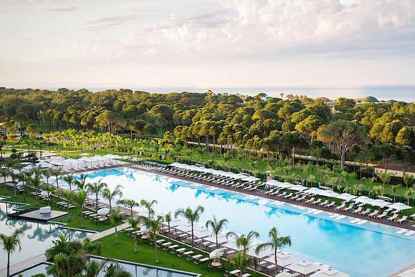 Außenansicht des Regnum Carya Golf & Spa Resorts mit großen Swimmingpools, Sonnenliegen und Sonnenschirmen.