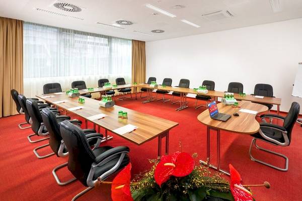 Konferenzraum im Star Inn Hotel Wien Schönbrunn mit langen Tischen, schwarzen Stühlen und roten Teppich.