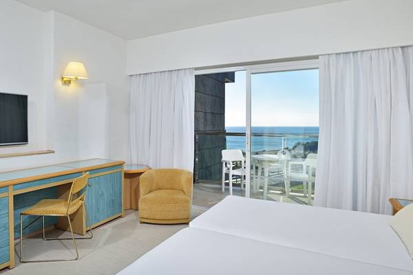 Innenansicht eines Hotelzimmers mit Meerblick, Doppelbett, Schreibtisch, Stühle und Balkon mit Tisch und Stühlen.