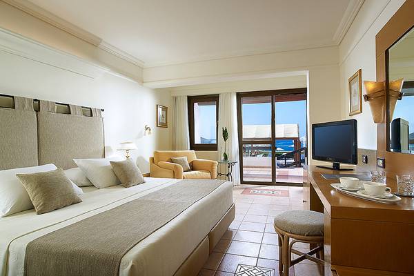 Hotelzimmer mit Doppelbett, Sessel, Fernseher und Balkon mit Meerblick.
