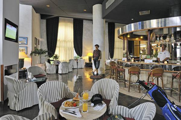 Hotellobby mit Bar und Golftasche: Gäste sitzen an Tischen, eine Bar mit Mann dahinter, Golftasche und Golfschläger neben dem Tisch.