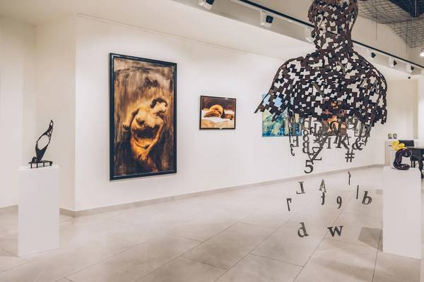 Ausstellung von Kunstwerken im Innenraum mit Gemälden an der Wand, einer Skulptur und einem großen Schriftbild.