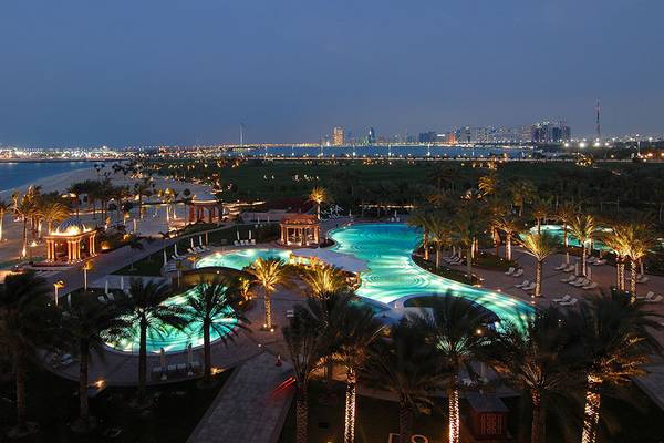 Abendliche Außenansicht des Emirates Palace Mandarin Oriental Hotels mit Swimmingpool, Palmen und Stadtsilhouette im Hintergrund.