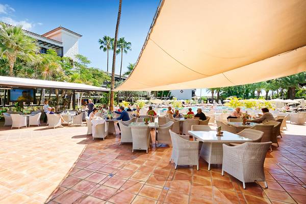 Terrasse des Parque Tropical Hotels mit Gästen und Sonnenschirmen, umgeben von Palmen und Gebäuden.