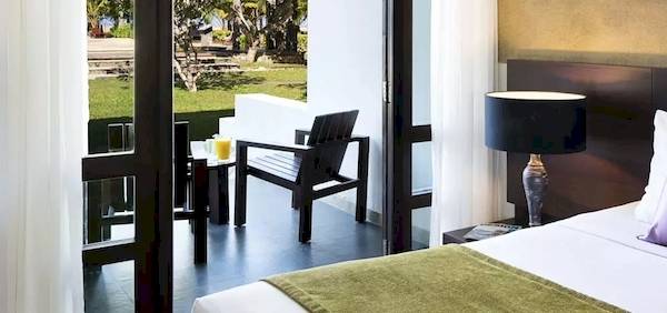 Hotelzimmer mit Balkon und Gartenblick, Bett, Kissen, Lampen und Tisch mit Getränken.