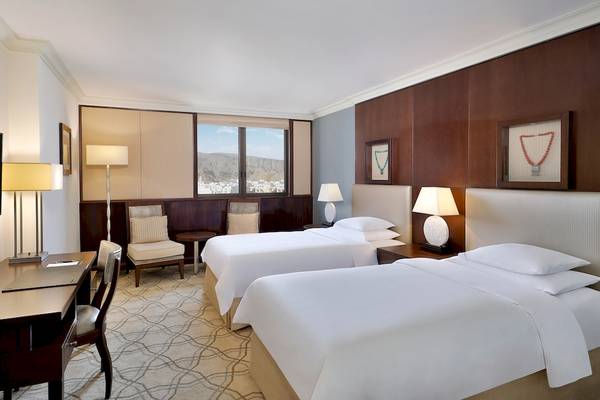Doppelzimmer im Sheraton Oman mit zwei Betten, Schreibtisch, Sesseln und Blick aus dem Fenster.