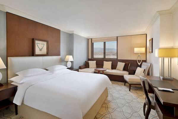 Schlafzimmer im Sheraton Oman Hotel mit Kingsize-Bett, Couch, Schreibtisch und Fenster mit Stadtblick.