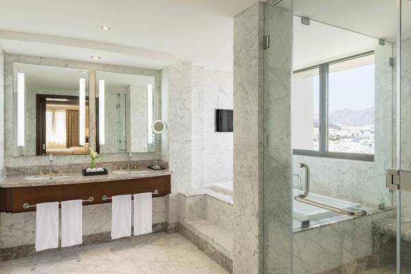 Badezimmer des Sheraton Oman Hotels mit Marmorfliesen, zwei Waschbecken, Spiegeln und einer Dusche.