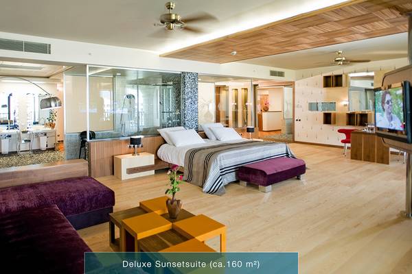 Innenansicht der Deluxe Sunsetsuite im Defne Kumul Deluxe Hotel mit Doppelbett, Fernseher, Wohnzimmerbereich und Glaswänden.