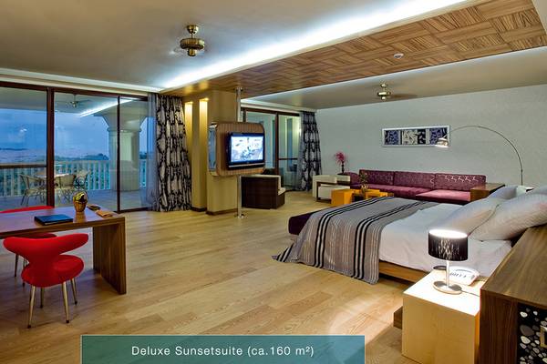 Innenansicht der Defne Kumul Deluxe Suite mit Balkon, Fernseher, Schreibtisch, rotem Stuhl und Doppelbett.