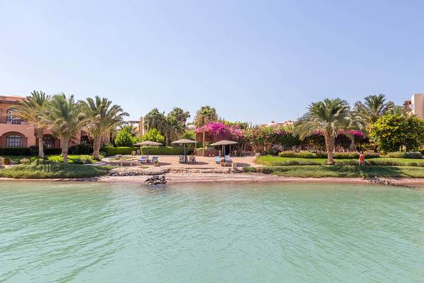Strandansicht des Sultan Bey Resorts mit Palmen, Liegestühlen und Gebäuden im Hintergrund.
