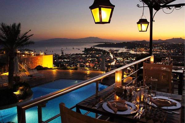Terrasse mit Aussicht auf den Hafen von Marmara Bodrum, mit beleuchtetem Schwimmbad, Tischen und Stühlen.