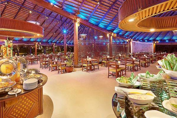 Innenansicht des Restaurants im Kuredu Island Resort & Spa mit vielen Tischen und Stühlen, Essensbuffets und blauen Beleuchtungselementen.