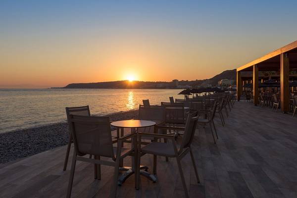 Terrasse des Avra Beach Resort Hotels mit Tischen und Stühlen am Meer bei Sonnenuntergang.