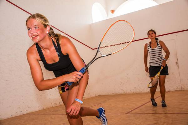 Zwei Frauen spielen Squash in einem Innenbereich mit Holzböden und weißen Wänden.