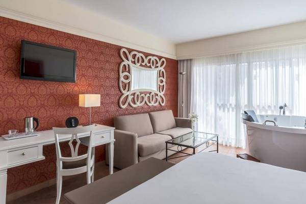 Luxuriöses Zimmer im Barcelo Corralejo Bay Hotel mit rotem Wanddesign, Fernseher, Schreibtisch, Couch, Badewanne und großer Fenster mit Vorhängen.