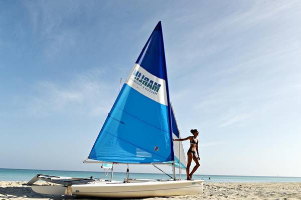 Eine Frau steht neben einem weißen Segelboot mit einer blauen Segel auf einem Strand im Royalton Hicacos Resort & Spa.
