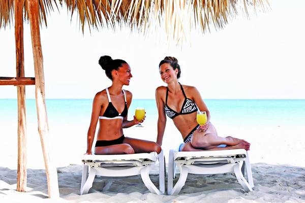Zwei Frauen auf Sonnenliegen am Strand unter einem Strohhut, beide lächeln und halten Gläser mit Getränken.