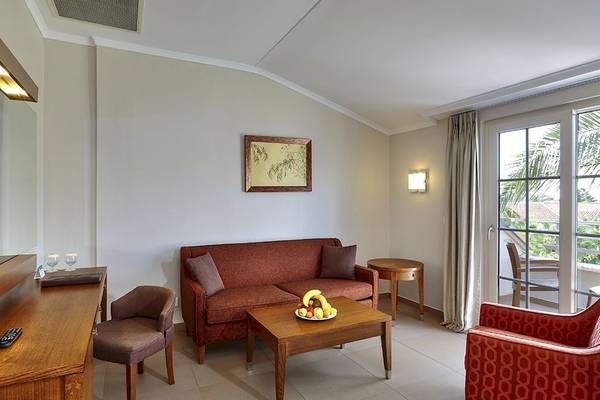 Wohnzimmer des Sunis Elita Beach Resort Hotel & Spa mit rotem Sofa, Tisch, Obstteller und Balkonzugang.