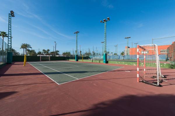 Tennisplatz im Freien mit Tor und Beleuchtungsmasten bei Sonnenschein.