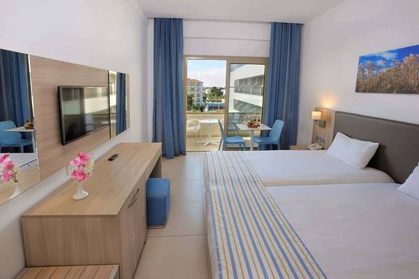 Zimmer im Nissiana Hotel mit Balkon und Meerblick, inklusive zwei Betten, Fernseher und Tisch mit Stühlen.